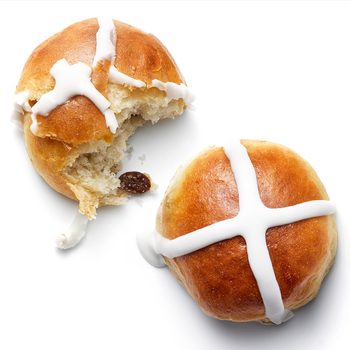 Buns with icing crosses rest on a plain white surface, one is partially eaten, revealing a raisin inside.