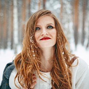 Woman wearing a white sweater, smiling slightly, stands in a snowy forest with snowflakes in her hair.