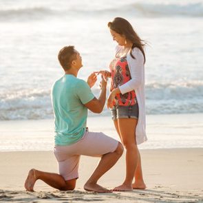 A man on one knee proposes on a sandy beach, offering a ring to a standing woman, with ocean waves in the background.