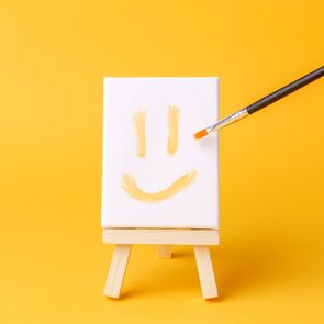 A small canvas displays a smiley face being painted with a brush, set on a wooden easel against an orange background.