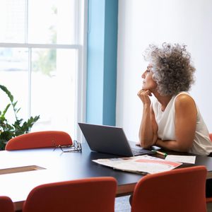 Woman sits contemplating, resting chin on hand, beside open laptop in a brightly lit office with red chairs and a potted plant near the window.