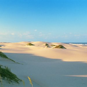 Sandy dunes stretch across the landscape, bordered by gentle ocean waves, under a clear blue sky.