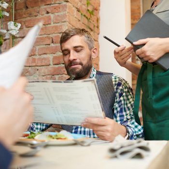 Portrait shot of bearded middle-aged man studying menu while unrecognizable waitress standing next to him and waiting for his order, interior of stylish restaurant on background