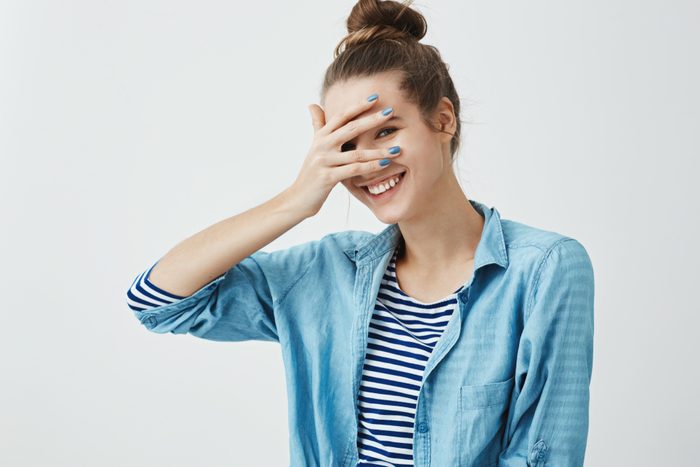 Girl saying silly words and gets embarrassed. Indoor shot of cute slender woman in stylish garment, covering eyes with hand but peeking, smiling broadly at camera, expressing positive emotions
