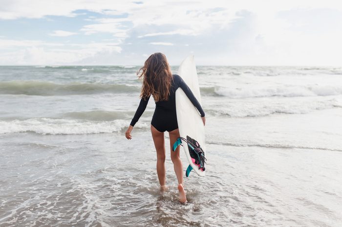 Young sporty active woman dressed swimsuit runs in the ocean with surf board in sunny day. Surfer girl walking with board on the sandy beach.