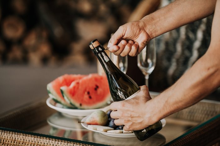Hands twist the muse on a bottle of sparkling wine. Back on the glass table a plate with watermelon and fruit