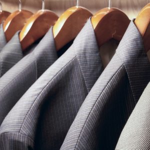 Jackets hang neatly on wooden hangers, arranged in a wardrobe with a variety of pinstripe patterns and subtle textures.