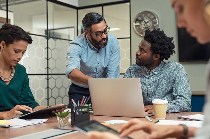 Mature manager discussing with his colleague online data in front of laptop. Serious businessman wearing eyeglasses training employees in job. Creative multiethnic team working together with boss in a meeting room.