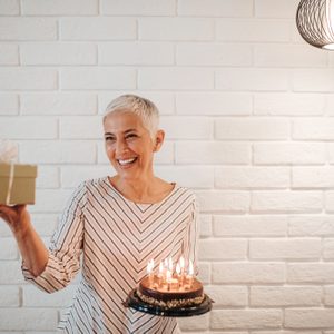 A smiling person holds a lit birthday cake and a gift, standing in front of a white brick wall under a modern pendant light.