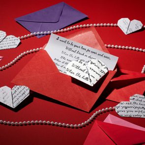Envelope holds a letter on a red surface, surrounded by origami heart shapes and pearl strings. Other colorful envelopes lie nearby.