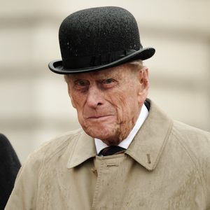 Elderly man wearing a black bowler hat and beige coat, standing outdoors. Background is blurred. Medals are partially visible on a person beside him.