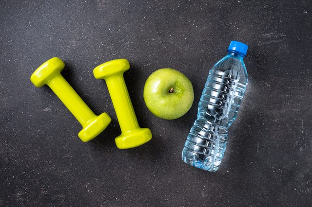 Dumbbells lie beside an apple and water bottle on a dark, textured surface.
