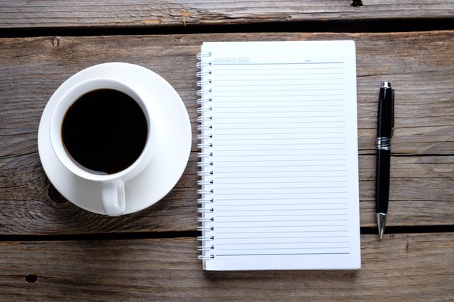 A cup holds coffee, resting beside a lined notebook and pen on a rustic wooden table.