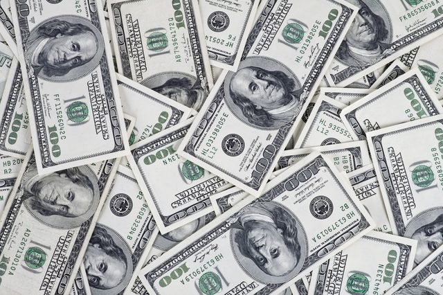 Hundred-dollar bills scattered, overlapping each other, creating a dense pile, primarily showing portraits and numerical values.