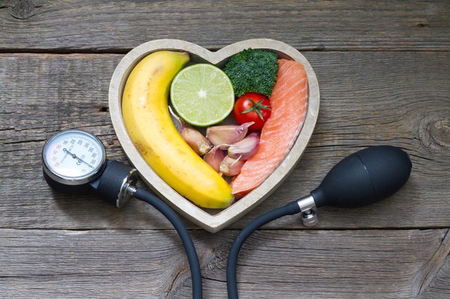 Heart-shaped bowl contains banana, lime, broccoli, tomato, garlic, and salmon. Blood pressure monitor rests beside on rustic wooden surface.