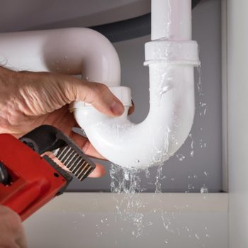 Pipe leaks water; a hand tightens it with a red wrench; located under a sink.