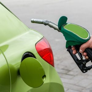Fuel pump being held, positioned near a green car's open fuel tank on a paved surface.
