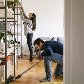 Woman reaches for shelf while man vacuums nearby; setting is a living room with plants and wooden shelves.