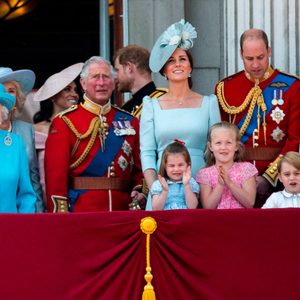 People stand smiling on a balcony, dressed in formal attire with medals. They appear to be at a public event, in front of a red draped railing.