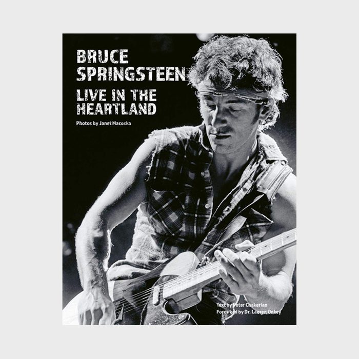 Musician plays guitar energetically on stage, wearing a sleeveless plaid shirt. Bold text reads 