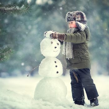 Child building snowman in snowy forest, wearing winter coat and hat, surrounded by snow-covered trees.