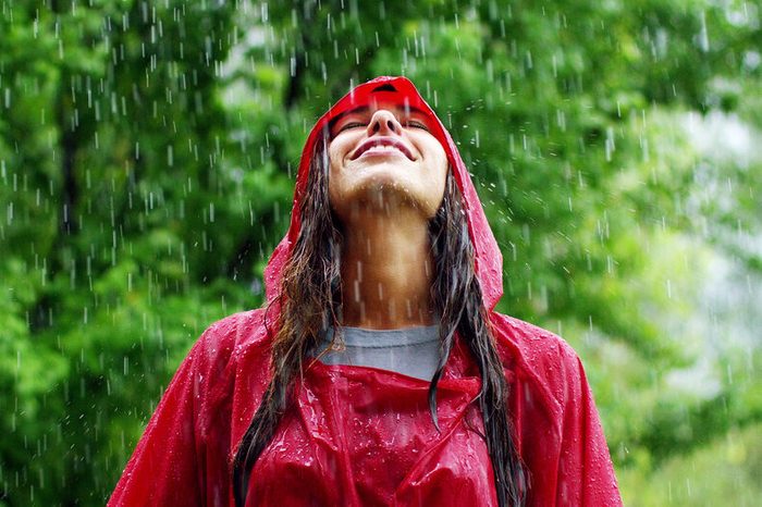 Person in red raincoat smiling, enjoying falling rain, surrounded by lush green trees.