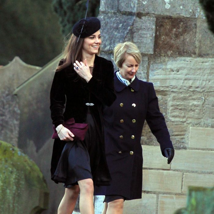 Two women walk past a stone wall, dressed formally, on a grassy area with headstones nearby.