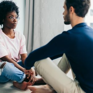 Two people sit cross-legged, engaged in conversation, in a bright room with a large window.