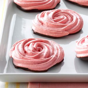 Pink meringue cookies shaped like roses rest on a white rectangular tray, with a chocolate base, atop a patterned tablecloth.