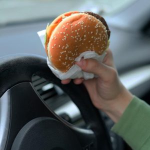 A hand holds a sesame seed hamburger inside a car, next to the steering wheel.