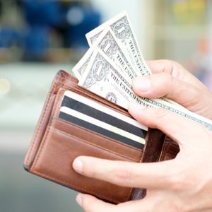 Hands hold a brown wallet containing credit cards and three one-dollar bills, set against a blurred indoor background.