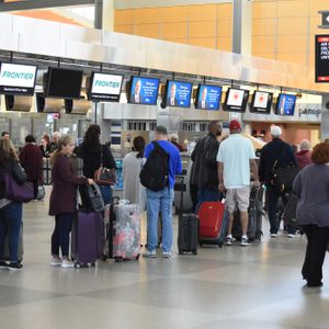 People wait in line at an airport check-in counter; luggage surrounds them, and airline screens display information above.