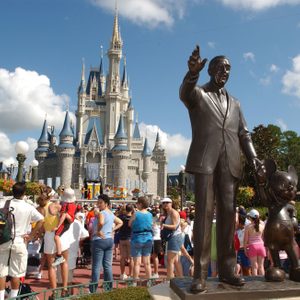 Bronze statue of a man and mouse holding hands, surrounded by tourists, set against a large fairytale castle, with a clear blue sky.