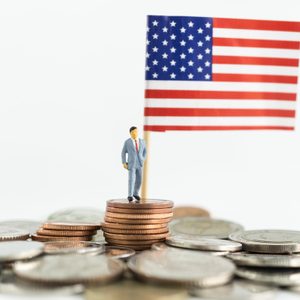 Miniature figure stands on stacked coins, holding a small American flag, amidst a pile of scattered coins.