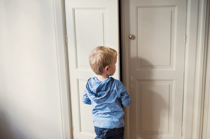 A rear view of toddler boy standing near door inside in a bedroom.