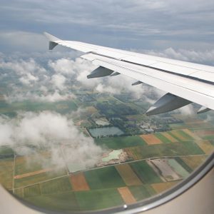 Airplane wing slices through the sky, viewed from window, above a patchwork of fields and scattered clouds.