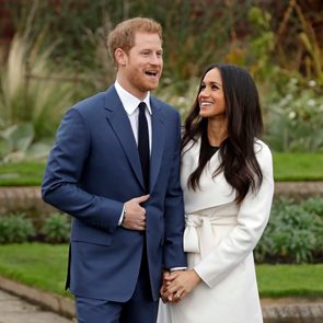 A man in a blue suit, smiling, stands beside a woman in a white coat; they hold hands in a garden setting.