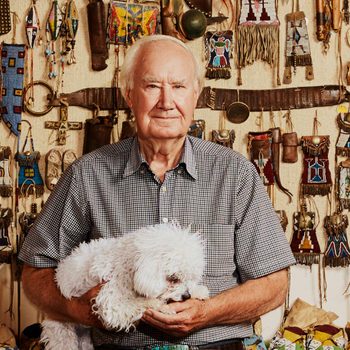 Elderly man holding a fluffy white dog, standing against a wall decorated with colorful Native American artifacts.