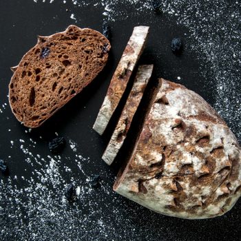 Sliced dark bread rests on a black, flour-dusted surface with scattered raisins.
