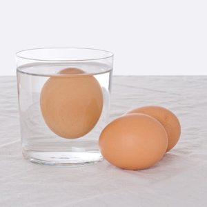 An egg floats in a glass of water; two eggs rest beside it on a white cloth surface.