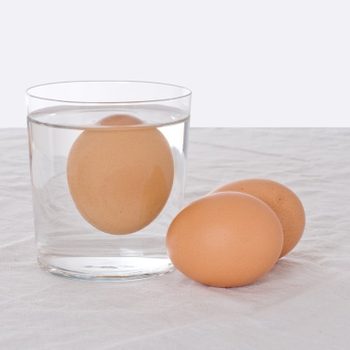 An egg floats in a glass of water; two eggs rest beside it on a white cloth surface.