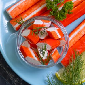 Crab sticks rest on a blue plate, garnished with dill and lemon slices, accompanied by a small dish of chopped pieces.