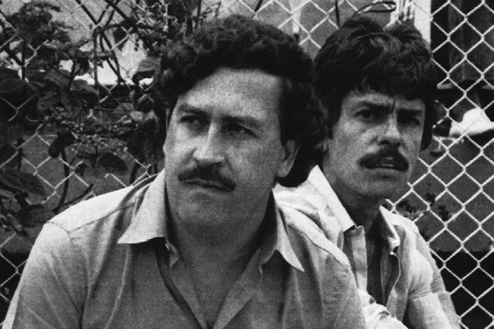 Two men sit beside a chain-link fence, one holding a cigarette, appearing contemplative in an outdoor setting.