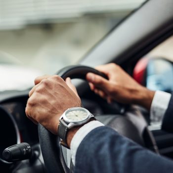Hands grip a car steering wheel, wearing a watch, inside a vehicle. A blurred exterior is visible through the window.