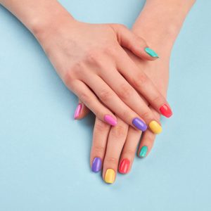 Hands rest on a light blue surface, displaying vibrantly colored nails in pink, purple, yellow, red, and teal.