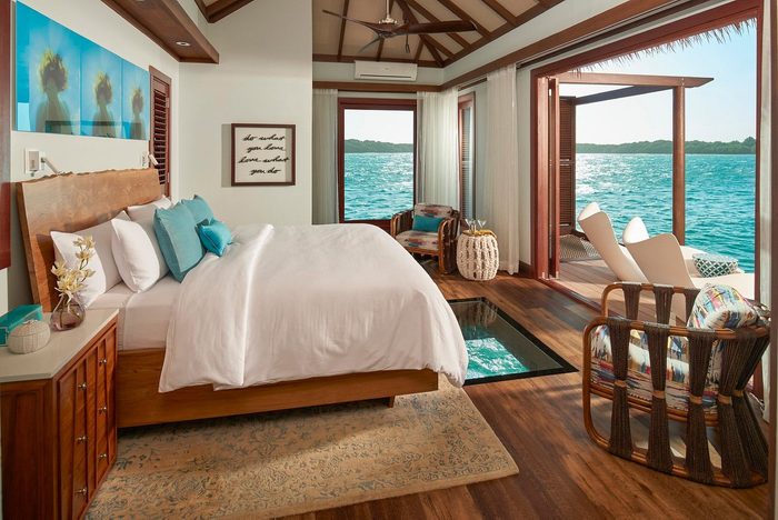 Bedroom overlooks ocean through open balcony, featuring elegant decor, cozy bed, and wooden furniture. Walls have abstract art and inspiring text: 