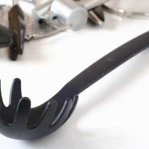 A black pasta server rests on a white surface, surrounded by various kitchen utensils in the background.
