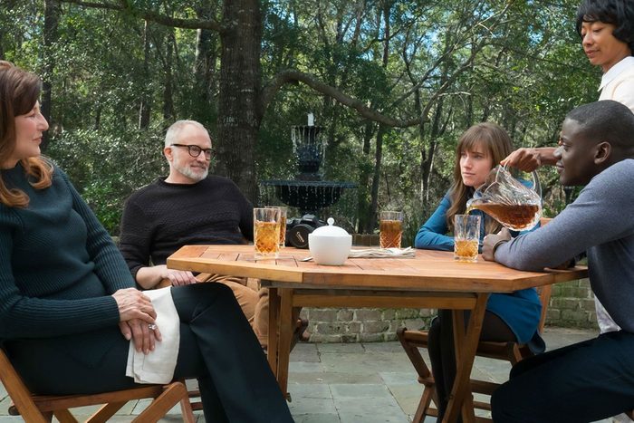 People sit around a wooden patio table with drinks, as one person pours tea from a glass pitcher, surrounded by trees.