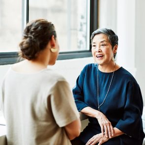 Two women engage in conversation near a window in a bright, modern office with a small plant on the table.