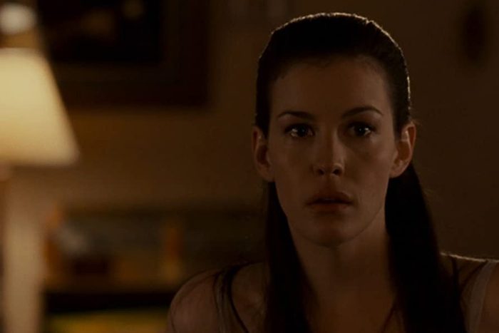 A woman with long dark hair appears startled, sitting in a dimly lit room with a lamp and blurred wall art in the background.